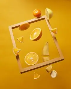 Composition artistique de fruits coupés — orange, banane, citron — flottant dans un cadre en bois sur fond jaune, symbolisant une alimentation vivante, colorée et adaptée au cycle féminin.